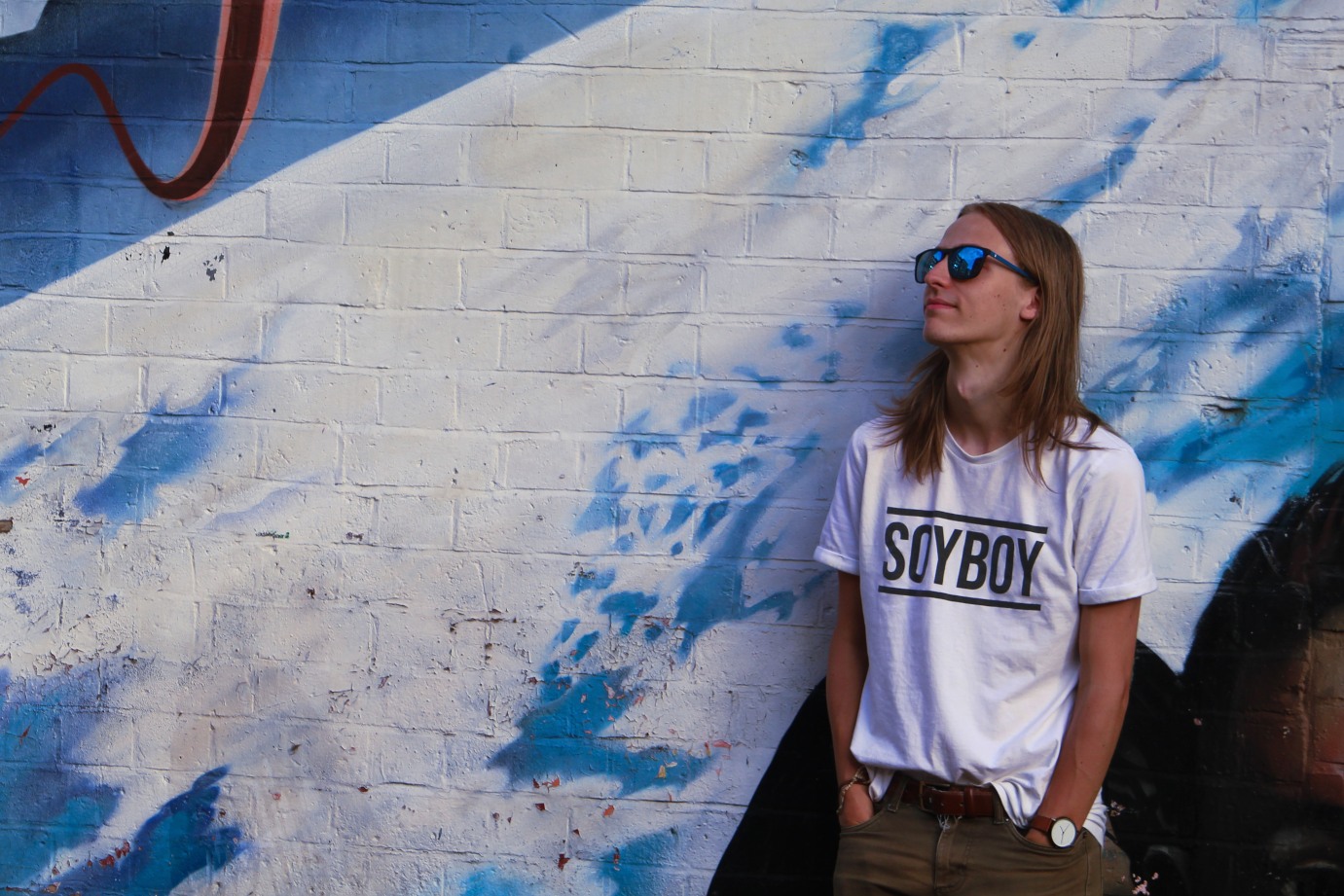 Portrait photo of a skinny white guy with long blond hair wearing sunglasses and a white t-shirt that says “SOYBOY” while stood against a blue and white mural trying his best to look cool